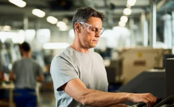 NR-1 atualizada reforça prevenção no ambiente de trabalho Trabalhador industrial usando óculos de proteção em máquina, em matéria sobre NR-1 e prevenção de riscos no ambiente de trabalho.