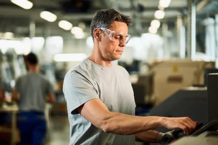 NR-1 atualizada reforça prevenção no ambiente de trabalho Trabalhador industrial usando óculos de proteção em máquina, em matéria sobre NR-1 e prevenção de riscos no ambiente de trabalho.