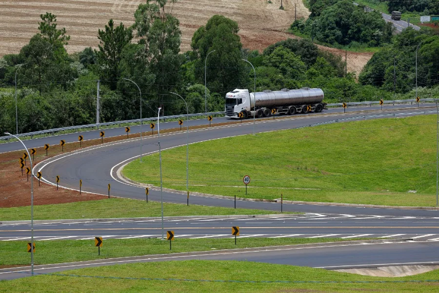 Caminhão tanque branco trafega em curva de rodovia estadual de Santa Catarina com asfalto novo, sinalização vertical revitalizada e defensas metálicas, ilustrando as melhorias do programa Estrada Boa na infraestrutura viária.