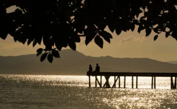 Santa Catarina terá calor de 32°C e queda de temperatura Silhueta de duas pessoas sentadas em um trapiche à beira-mar durante o pôr do sol em Santa Catarina. O reflexo do sol brilha na água, ilustrando o tempo firme e as temperaturas elevadas de outono no estado.