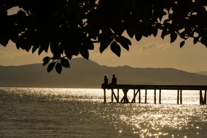Santa Catarina terá calor de 32°C e queda de temperatura Silhueta de duas pessoas sentadas em um trapiche à beira-mar durante o pôr do sol em Santa Catarina. O reflexo do sol brilha na água, ilustrando o tempo firme e as temperaturas elevadas de outono no estado.