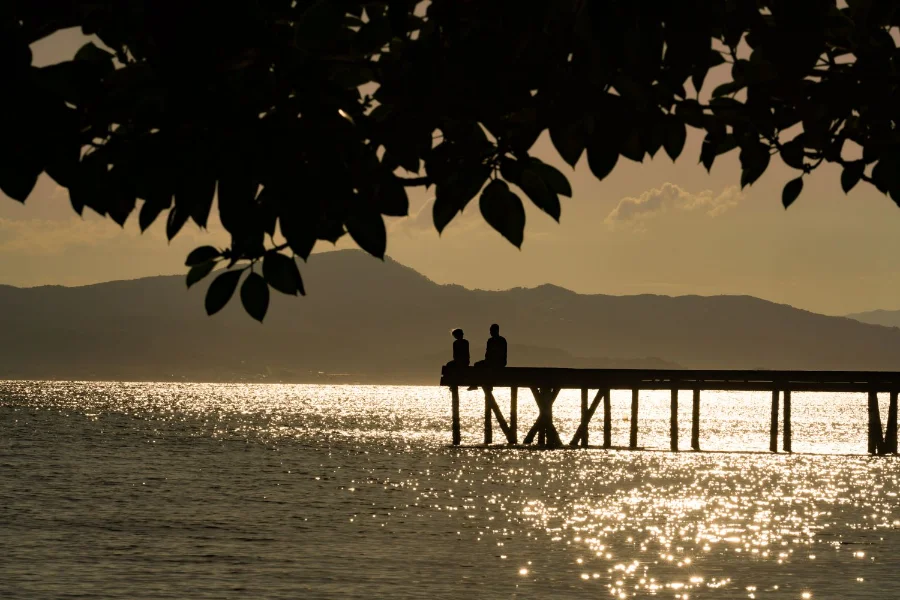 Silhueta de duas pessoas sentadas em um trapiche à beira-mar durante o pôr do sol em Santa Catarina. O reflexo do sol brilha na água, ilustrando o tempo firme e as temperaturas elevadas de outono no estado.