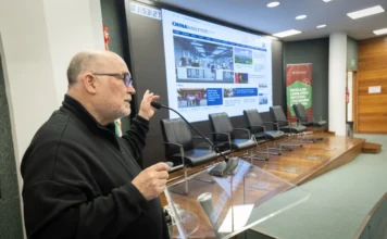 Alesc realiza seminário sobre evolução da educação na China O palestrante e pesquisador Milton Pomar fala ao microfone no púlpito durante o seminário sobre educação na China na Alesc. Ao fundo, uma grande tela exibe o site China Daily em um auditório com cadeiras vazias no palco.