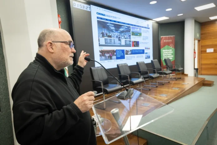 Alesc realiza seminário sobre evolução da educação na China O palestrante e pesquisador Milton Pomar fala ao microfone no púlpito durante o seminário sobre educação na China na Alesc. Ao fundo, uma grande tela exibe o site China Daily em um auditório com cadeiras vazias no palco.