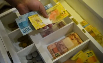 Caixa conclui Bolsa Família de junho para NIS final 0 Mão segurando notas de dinheiro em gaveta de caixa, em imagem relacionada ao pagamento do Bolsa Família de junho para beneficiários com NIS final 0.