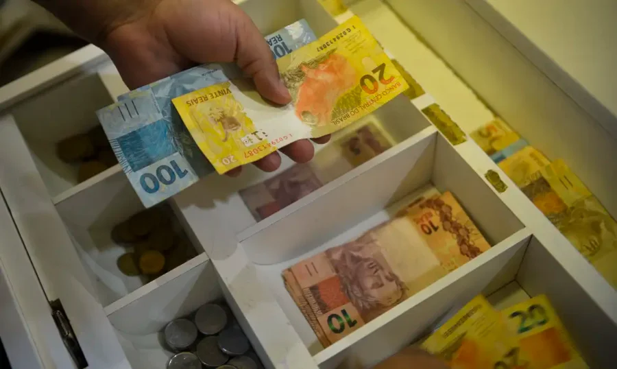 Mão segurando notas de dinheiro em gaveta de caixa, em imagem relacionada ao pagamento do Bolsa Família de junho para beneficiários com NIS final 0.