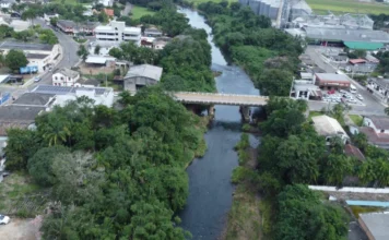 Jacinto Machado planeja prevenção contra chuvas do El Niño Vista aérea do Rio Pinheirinho em Jacinto Machado, com ponte, área urbana e vegetação em região monitorada para prevenção contra enxurradas do El Niño.