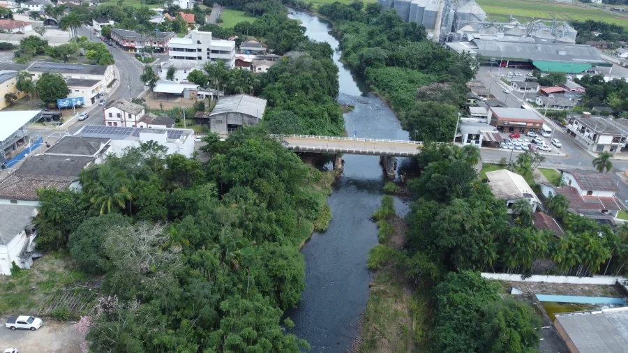 Vista aérea do Rio Pinheirinho em Jacinto Machado, com ponte, área urbana e vegetação em região monitorada para prevenção contra enxurradas do El Niño.