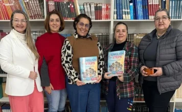 Araranguá capacita professores com novo material de inglês Cinco educadoras de Araranguá posam em uma biblioteca segurando exemplares do novo material didático "English Steps" para turmas do Ensino Fundamental, focando em formação de professores e BNCC.