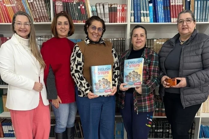 Araranguá capacita professores com novo material de inglês Cinco educadoras de Araranguá posam em uma biblioteca segurando exemplares do novo material didático "English Steps" para turmas do Ensino Fundamental, focando em formação de professores e BNCC.
