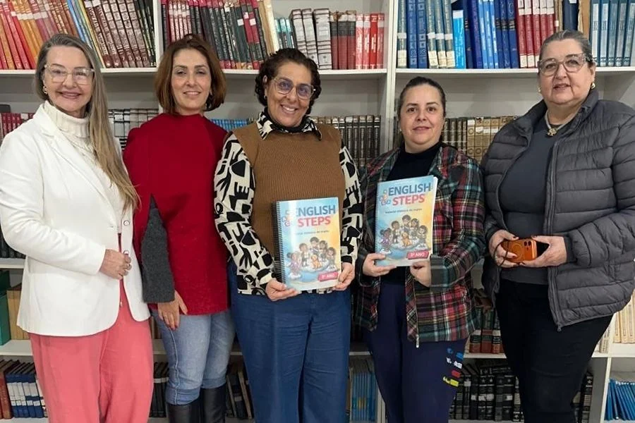 Cinco educadoras de Araranguá posam em uma biblioteca segurando exemplares do novo material didático 