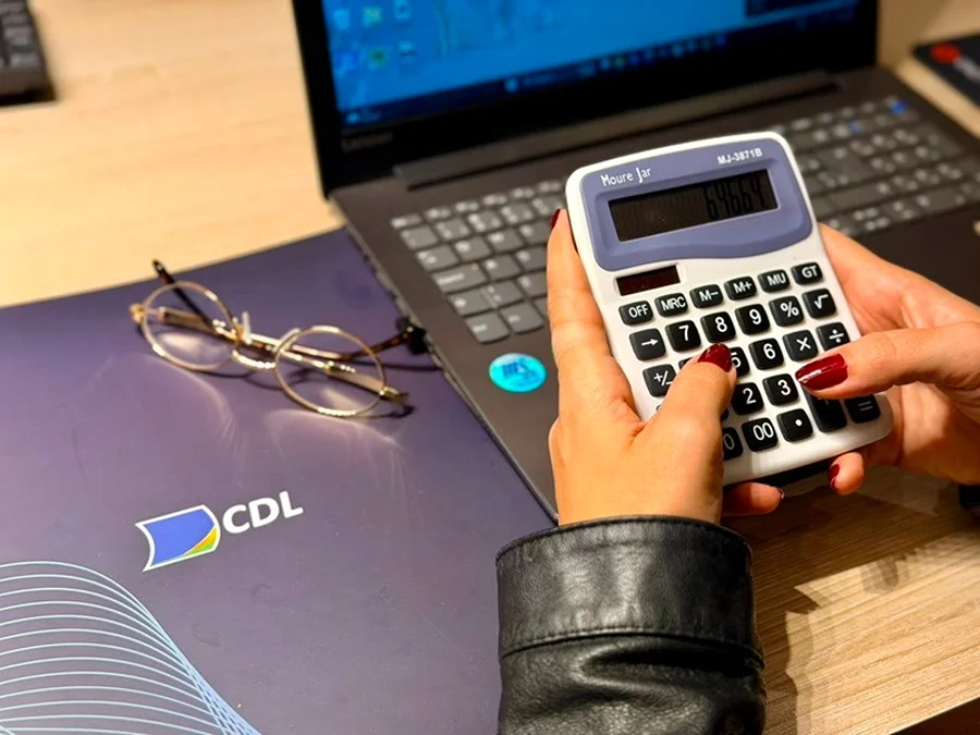 Mãos utilizam calculadora sobre mesa com notebook e pasta da CDL, ilustrando oficina gratuita de precificação para empreendedores em Araranguá.