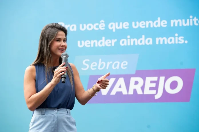 CDL Criciúma realiza workshop sobre vendas em alta performance Fabrícia Borba, especialista em marketing e vendas e consultora do Sebrae, palestranda com um microfone na mão em um evento de capacitação para o varejo da CDL Criciúma. O fundo é azul com texto de apoio.