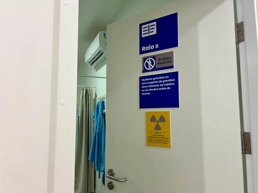 Equipamento de raio-x completa um ano e ultrapassa 5,6 mil laudos em Siderópolis Porta branca da sala de raio-x na Policlínica São Lucas em Siderópolis, com placas de sinalização azul e amarela indicando acesso restrito e avisos de segurança sobre radiação e gravidez.