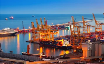 FIESC lidera missão empresarial à Expo Paraguay-Brasil 2026 Vista aérea de um porto comercial movimentado e iluminado ao anoitecer, destacando navios cargueiros, grandes guindastes amarelos e pilhas de contêineres. A infraestrutura logística ilustra o setor de comércio exterior, a exportação e a internacionalização da indústria catarinense, temas centrais da missão empresarial da FIESC na Expo Paraguay-Brasil.