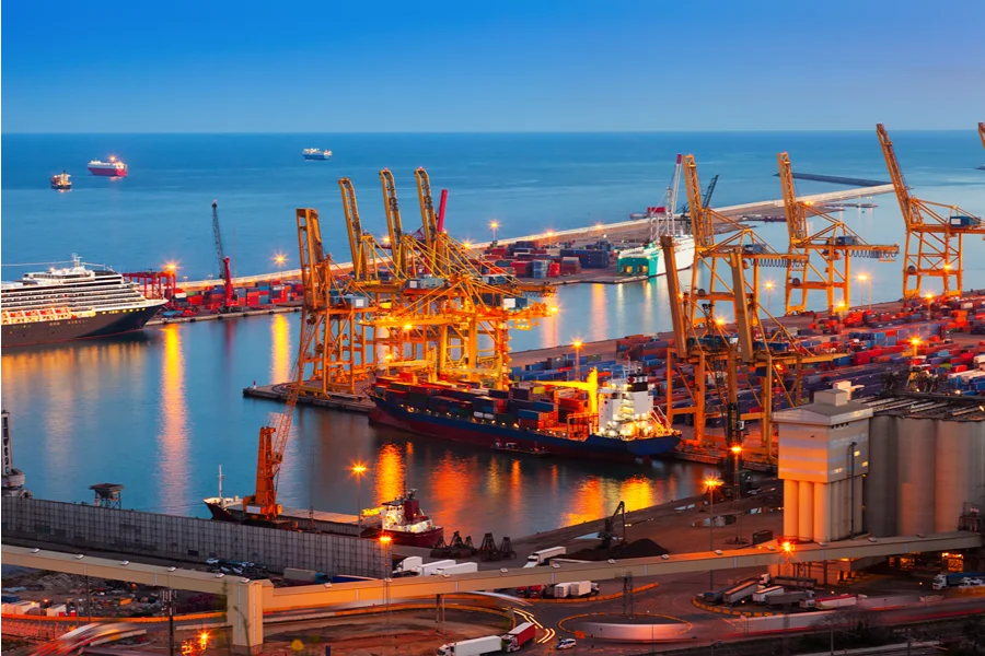 FIESC lidera missão empresarial à Expo Paraguay-Brasil 2026 Vista aérea de um porto comercial movimentado e iluminado ao anoitecer, destacando navios cargueiros, grandes guindastes amarelos e pilhas de contêineres. A infraestrutura logística ilustra o setor de comércio exterior, a exportação e a internacionalização da indústria catarinense, temas centrais da missão empresarial da FIESC na Expo Paraguay-Brasil.