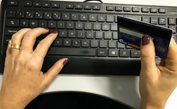 Governo regulamenta e-commerce em compras públicas Pessoa segura cartão bancário enquanto usa teclado de computador, em imagem que ilustra comércio eletrônico e compras públicas por plataformas digitais.