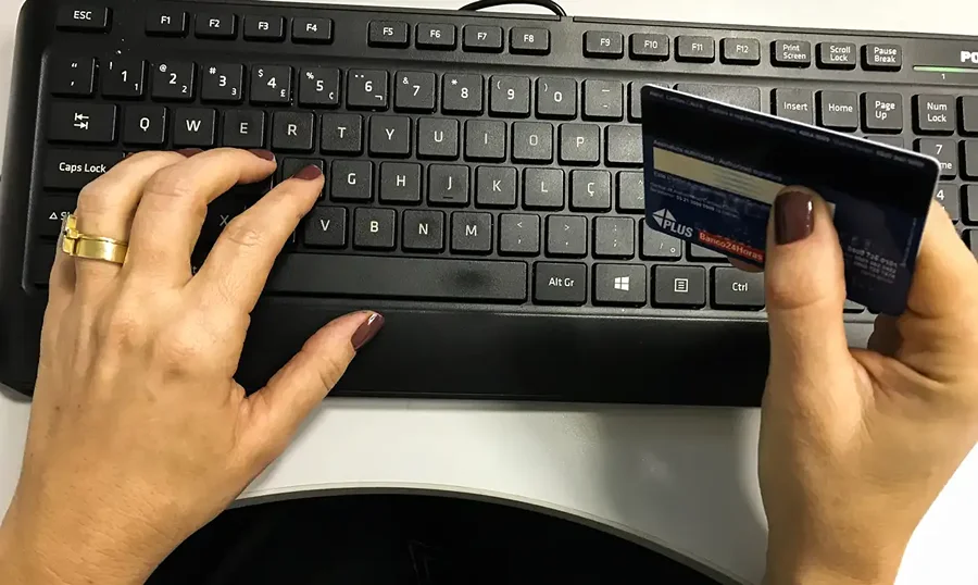 Pessoa segura cartão bancário enquanto usa teclado de computador, em imagem que ilustra comércio eletrônico e compras públicas por plataformas digitais.