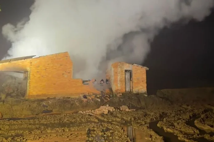 Incêndio destrói dois depósitos de madeira em Jacinto Machado Edificação destruída por incêndio em Jacinto Machado, com intensa fumaça branca saindo das ruínas de tijolos durante a noite. O terreno agrícola e enlameado é iluminado pelos refletores do Corpo de Bombeiros durante o trabalho de rescaldo.