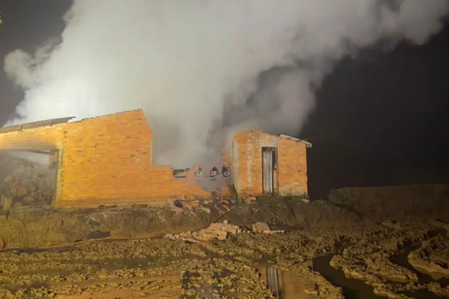 Edificação destruída por incêndio em Jacinto Machado, com intensa fumaça branca saindo das ruínas de tijolos durante a noite. O terreno agrícola e enlameado é iluminado pelos refletores do Corpo de Bombeiros durante o trabalho de rescaldo.