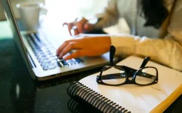 Mulheres são maioria nos seguros, mas minoria na liderança Mulher utiliza notebook ao lado de caderno e óculos, em imagem relacionada à participação feminina no mercado de seguros.
