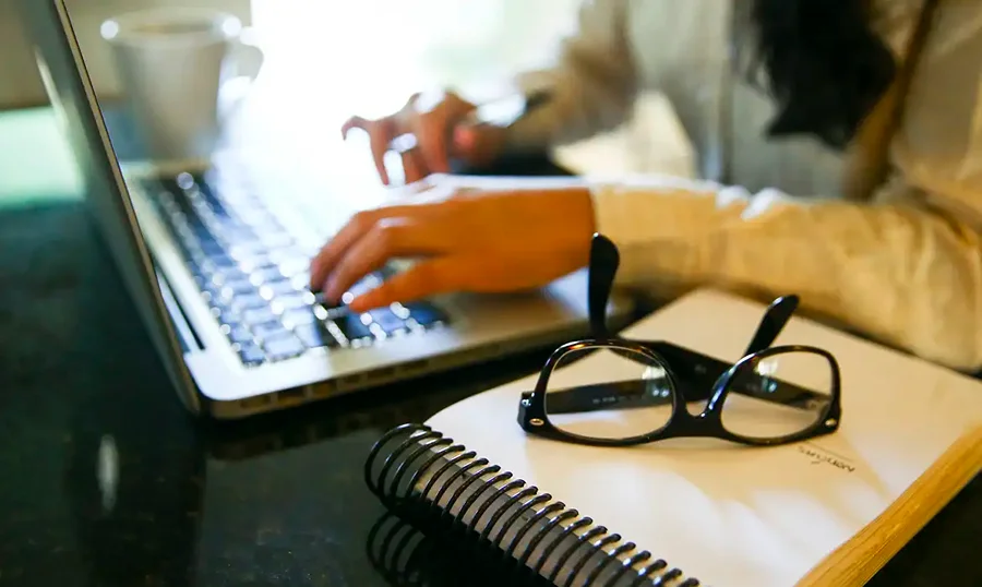 Mulher utiliza notebook ao lado de caderno e óculos, em imagem relacionada à participação feminina no mercado de seguros.