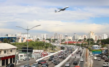 PNL 2050 prevê R$ 1,2 tri para logística brasileira Avião sobrevoa avenida com tráfego intenso em São Paulo, representando a integração entre transporte aéreo e rodoviário prevista no PNL 2050.