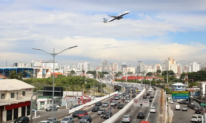 PNL 2050 prevê R$ 1,2 tri para logística brasileira Avião sobrevoa avenida com tráfego intenso em São Paulo, representando a integração entre transporte aéreo e rodoviário prevista no PNL 2050.