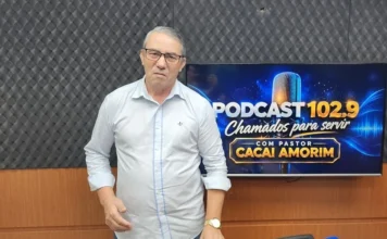 Pastor relata milagre de infância em programa na rádio 102.9 Pastor Reinaldo Martins, convidado do podcast Chamados para Servir na rádio 102.9 FM, de pé no estúdio, usando óculos e camisa branca, em frente a uma TV que exibe o logotipo do programa e o nome do apresentador.