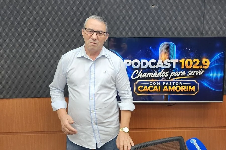 Pastor relata milagre de infância em programa na rádio 102.9 Pastor Reinaldo Martins, convidado do podcast Chamados para Servir na rádio 102.9 FM, de pé no estúdio, usando óculos e camisa branca, em frente a uma TV que exibe o logotipo do programa e o nome do apresentador.