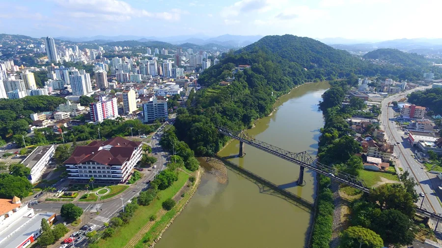 Vista aérea de Blumenau (SC), mostrando o centro urbano, o Rio Itajaí-Açu e a histórica Ponte de Ferro (Ponte Aldo Pereira de Andrade) em direção ao morro arborizado.