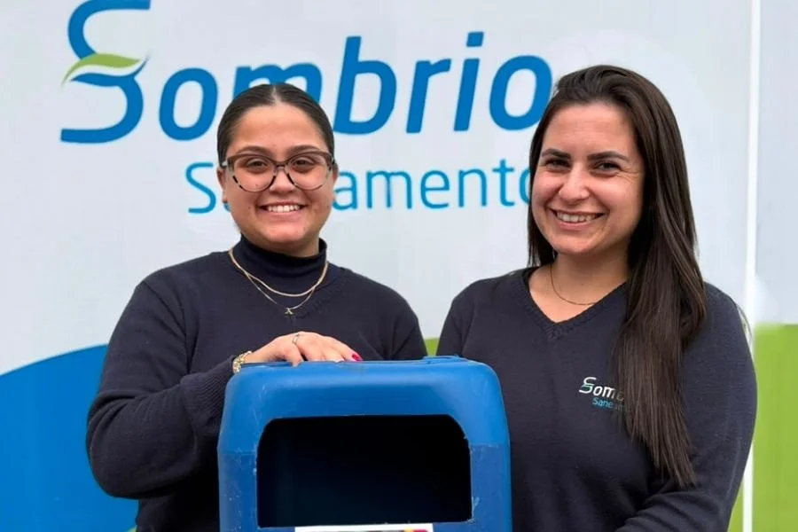 Duas colaboradoras da Sombrio Saneamento ao lado de coletor de tampinhas plásticas da campanha em apoio às APAEs de Sombrio e Balneário Gaivota.