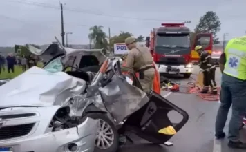 Homem é resgatado de ferragens após acidente em Criciúma Carro prata totalmente destruído em um acidente grave em Criciúma. Um bombeiro fardado está ao lado do veículo amassado realizando o resgate. Ao fundo, um caminhão de bombeiros e profissionais do SAMU atendem à ocorrência em uma rodovia.