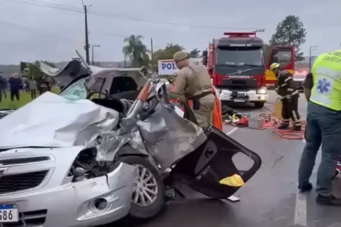 recorte-acidente-900x600 Carro prata totalmente destruído em um acidente grave em Criciúma. Um bombeiro fardado está ao lado do veículo amassado realizando o resgate. Ao fundo, um caminhão de bombeiros e profissionais do SAMU atendem à ocorrência em uma rodovia.