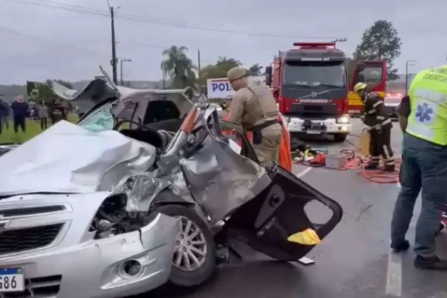 Carro prata totalmente destruído em um acidente grave em Criciúma. Um bombeiro fardado está ao lado do veículo amassado realizando o resgate. Ao fundo, um caminhão de bombeiros e profissionais do SAMU atendem à ocorrência em uma rodovia.