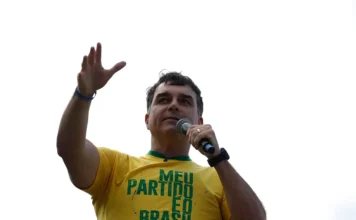 Aliado de Trump diz que Flávio Bolsonaro está nos EUA Flávio Bolsonaro fala ao microfone e acena com a mão, vestindo camiseta amarela.