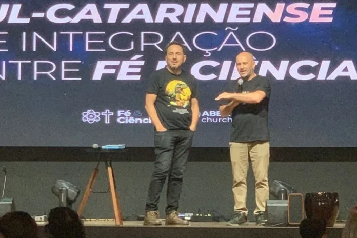 Criciúma terá simpósio gratuito para debater fé e ciência Dois homens no palco, um deles com microfone, diante de painel do Simpósio Sul Catarinense de Integração entre Fé e Ciência, em Criciúma.