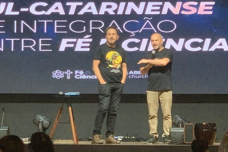 Criciúma terá simpósio gratuito para debater fé e ciência Dois homens no palco, um deles com microfone, diante de painel do Simpósio Sul Catarinense de Integração entre Fé e Ciência, em Criciúma.