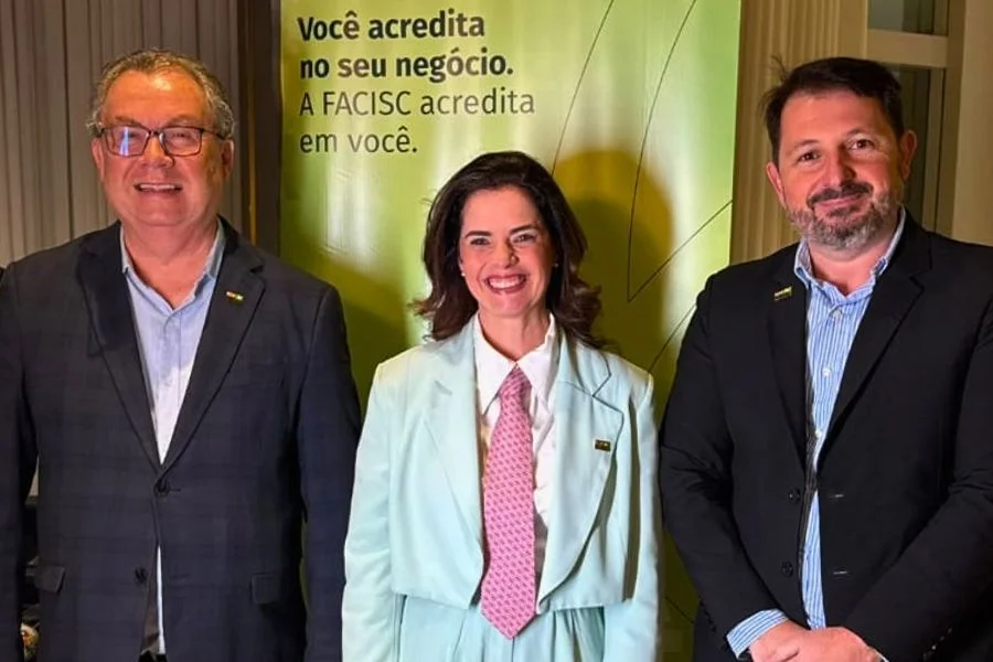 Três representantes do setor empresarial posam diante de um painel da FACISC.