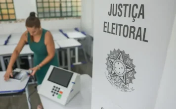 TSE registra troca de 900 urnas até o meio-dia no país Urna eletrônica ao lado de cabine da Justiça Eleitoral, com mulher ao fundo em seção de votação.