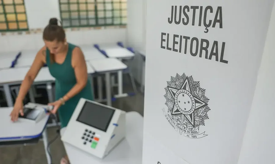 TSE registra troca de 900 urnas até o meio-dia no país Urna eletrônica ao lado de cabine da Justiça Eleitoral, com mulher ao fundo em seção de votação.
