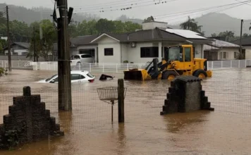 Timbé do Sul anuncia saque do FGTS após fortes chuvas Rua alagada em Timbé do Sul, com carro parcialmente submerso e máquina carregadeira diante de residências após fortes chuvas.