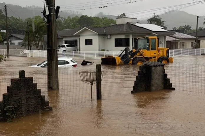 Timbé do Sul anuncia saque do FGTS após fortes chuvas Rua alagada em Timbé do Sul, com carro parcialmente submerso e máquina carregadeira diante de residências após fortes chuvas.