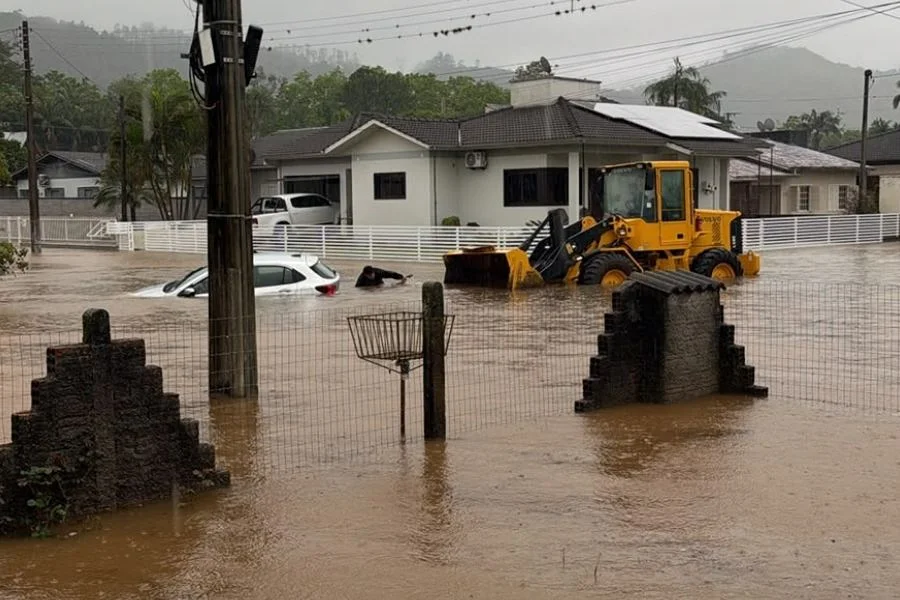 Timbé do Sul anuncia saque do FGTS após fortes chuvas Rua alagada em Timbé do Sul, com carro parcialmente submerso e máquina carregadeira diante de residências após fortes chuvas.