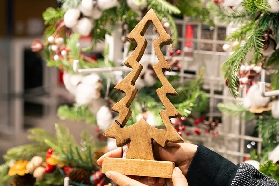 Mãos seguram árvore de Natal de madeira diante de enfeites natalinos, em imagem que ilustra o concurso de vitrines da CDL Araranguá.