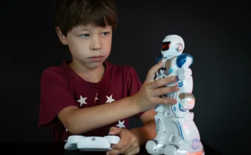 Brinquedos com IA: pediatras alertam para riscos às crianças Menino segura um controle remoto e toca um robô de brinquedo branco e azul sobre uma mesa.
