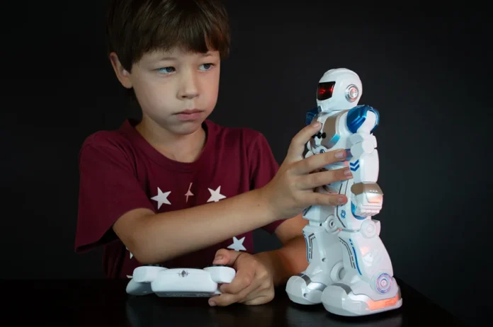 sunriseforever-boy-6480750 Menino segura um controle remoto e toca um robô de brinquedo branco e azul sobre uma mesa.