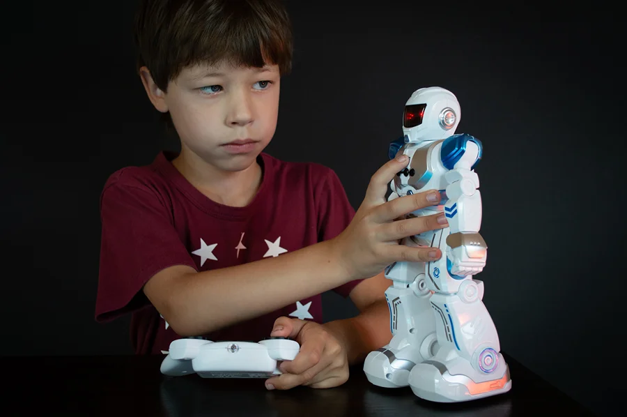 Menino segura um controle remoto e toca um robô de brinquedo branco e azul sobre uma mesa.